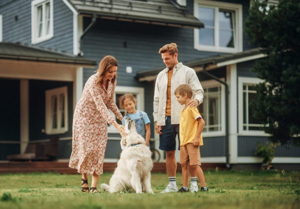 A woman, man, two children, and a white dog stand together on the lawn in front of a modern house. The scene evokes family unity—something an Akron property division lawyer understands during times of change. The woman is petting the dog while the others watch.